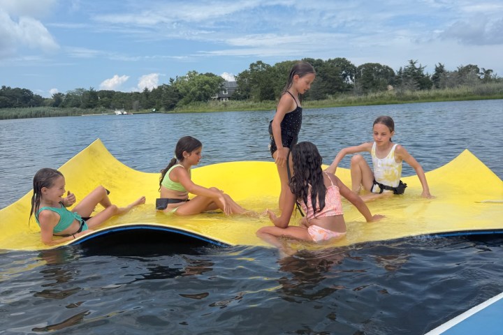 Five children playing on a yellow floating mat in the water under a blue sky.
