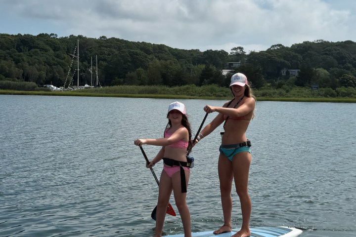 Two people paddleboarding on calm water under a partly cloudy sky, wearing swimwear and pink caps.