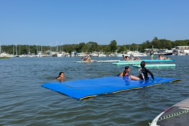 People on a blue floating mat and swimming in a lake with boats in the distance.