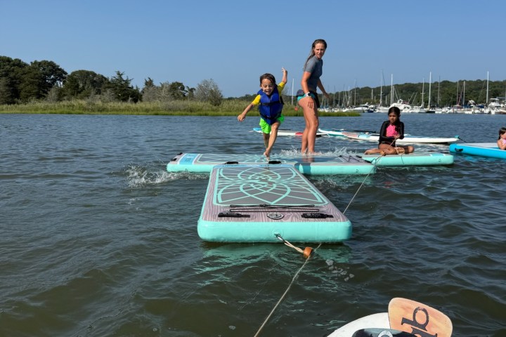 Child jumping off paddleboard into water beside two seated people on boards.