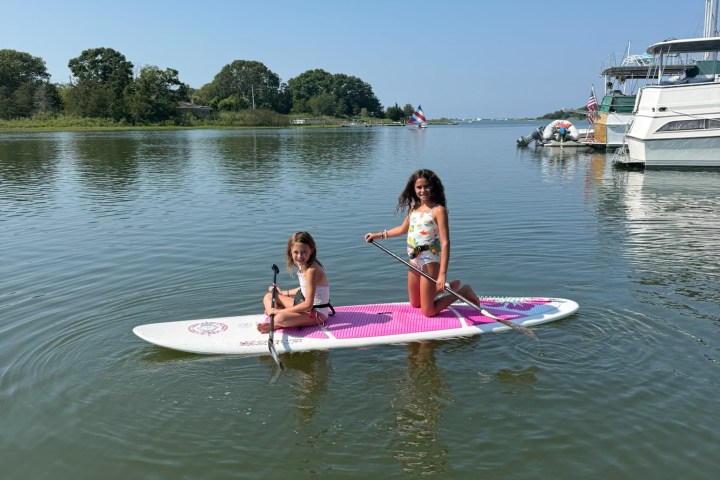 Two girls paddleboarding on a calm lake with boats and trees in the background.