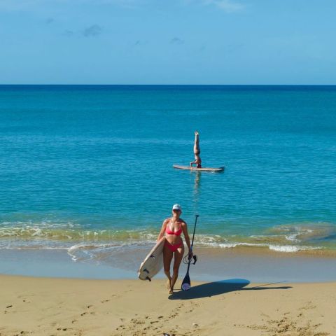 Person in red bikini holding paddleboard on beach; another doing headstand on board in calm sea.