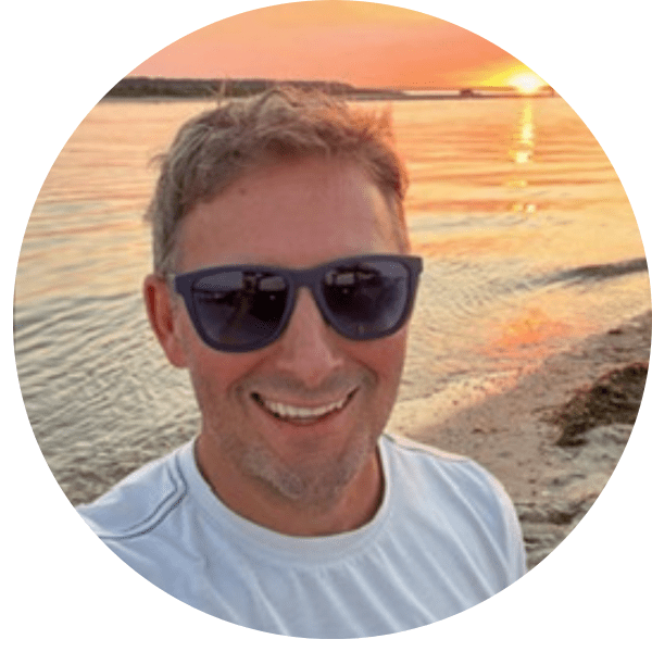 Man with sunglasses smiles at sunset on a beach.