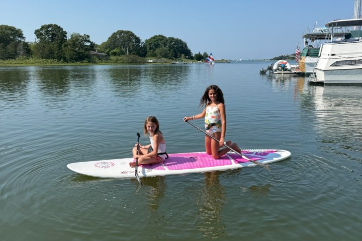 Two girls on a pink paddleboard in calm water with boats in the background.