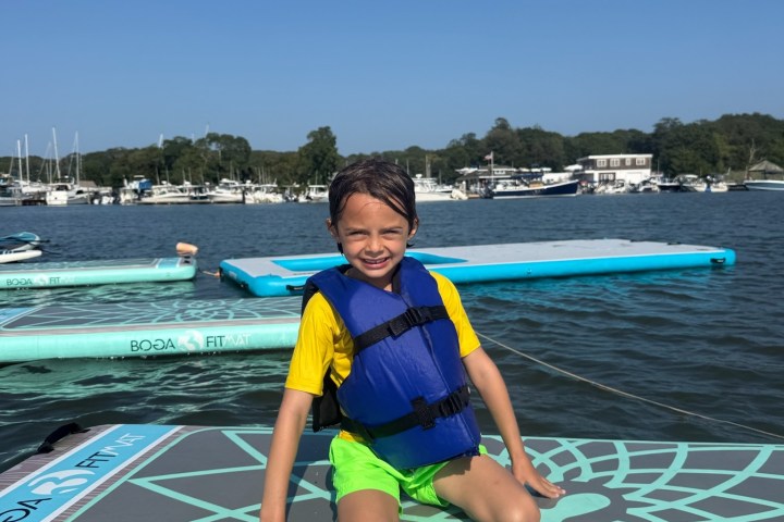 Child in life jacket kneeling on a paddleboard on a sunny day with boats and trees in the background.